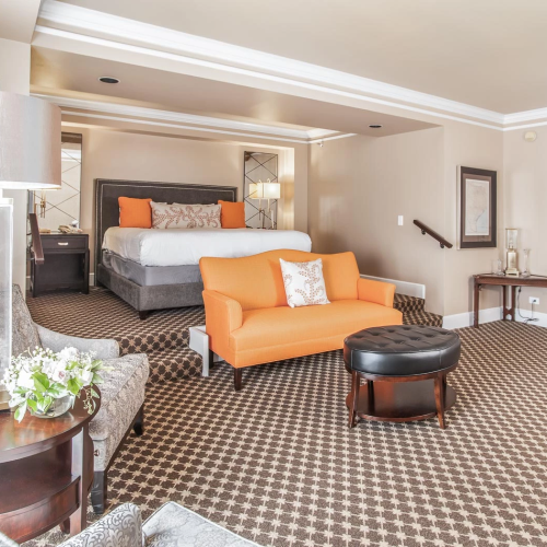 A spacious hotel suite with a bed, orange sofa, round ottoman, patterned carpet, and modern seating area under soft lighting, in neutral tones. End sentence.
