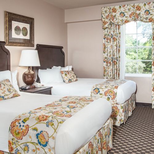 Two twin beds with white linens, floral bed runners, matching curtains, a nightstand lamp, and a window view in a cozy hotel room.
