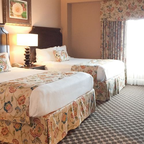 Two double beds with floral bed skirts and pillows, a nightstand lamp between them, a framed picture above, and a bright window with floral drapes.