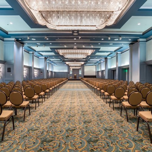 A grand conference hall with rows of beige chairs facing a long stage, ornate chandeliers, and a patterned carpet, set for a formal event.