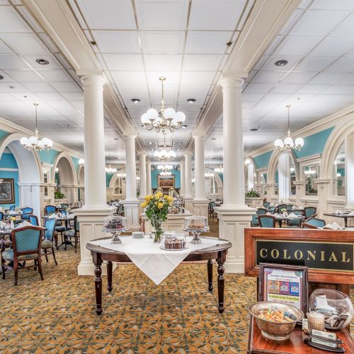 A bright, ornate classroom with high ceilings, chandeliers, rows of desks, and a long central table, feeling like a grand library or museum study hall.