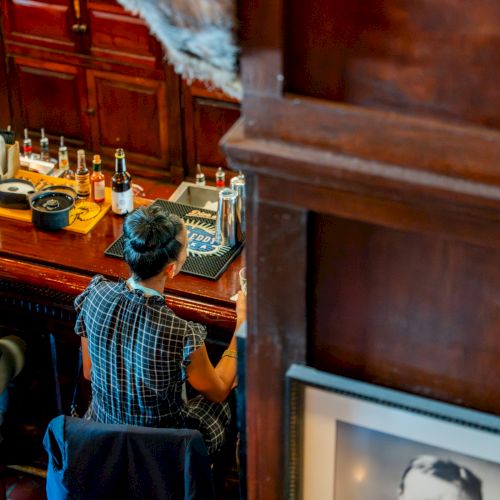 A man wearing a cap hands a drink to another in a cozy, wood-paneled bar, near glasses on a shelf.