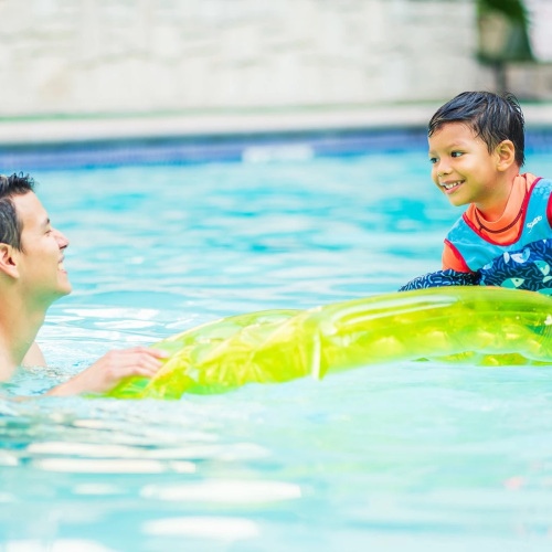 Two people in a pool: an adult man in the water and a smiling child on an inflatable raft, enjoying a sunny swim together.