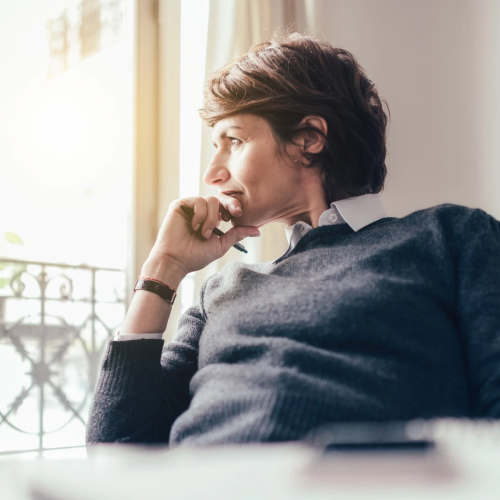 A man sits pensively by a window, resting his chin on his hand, wearing a gray sweater; soft daylight filters in, creating a calm, contemplative scene.