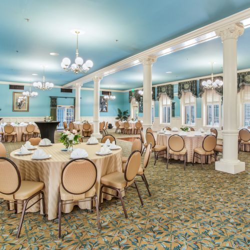 A bright, elegant banquet hall with blue ceilings, round tables dressed in beige cloths, and cream chairs, supported by white columns.