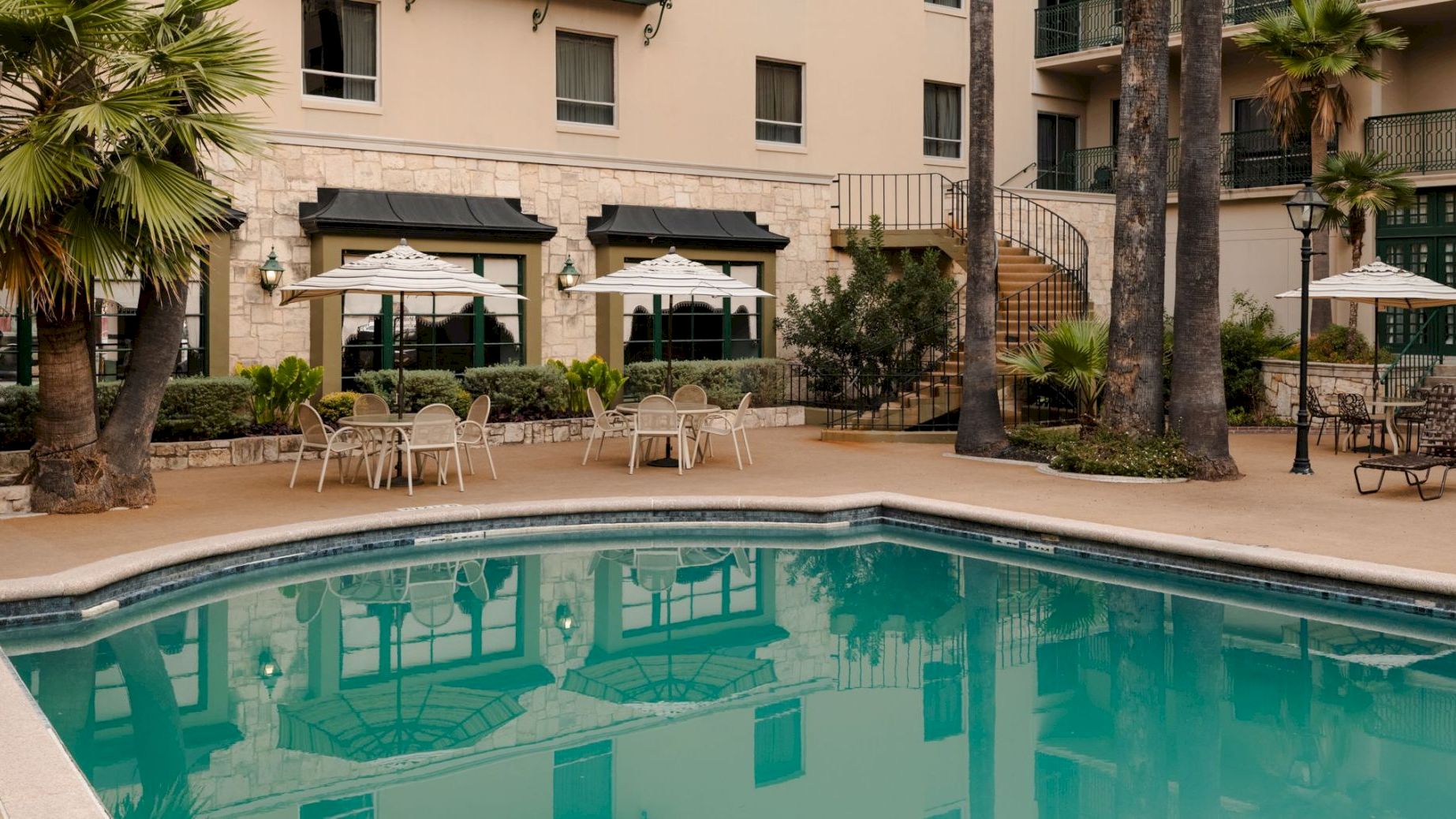 Courtyard pool with lounge chairs under striped umbrellas, palm trees, and a multi-story hotel building in view.