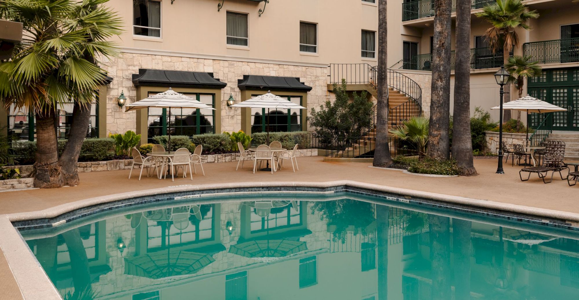 An outdoor hotel-style pool area with turquoise water, palm trees, patio tables with umbrellas, and a beige multi-story building in the background.