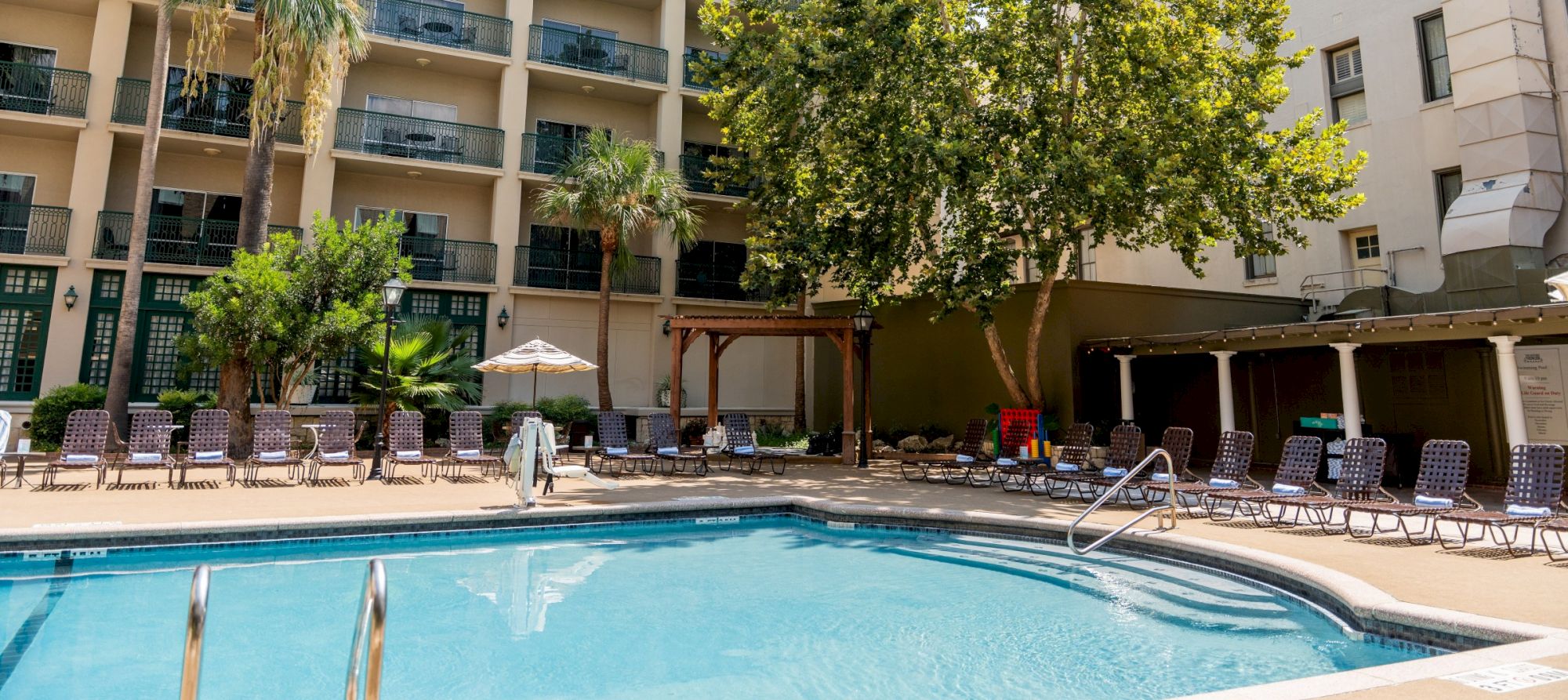 A hotel pool area with lounge chairs, palm trees, and a building backdrop under sunny skies, perfect for a relaxing day by the water.