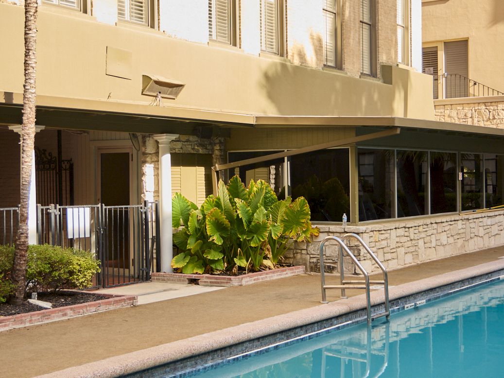 A sunny hotel courtyard with a blue swimming pool and beige buildings, featuring multiple green awning balconies and windows.