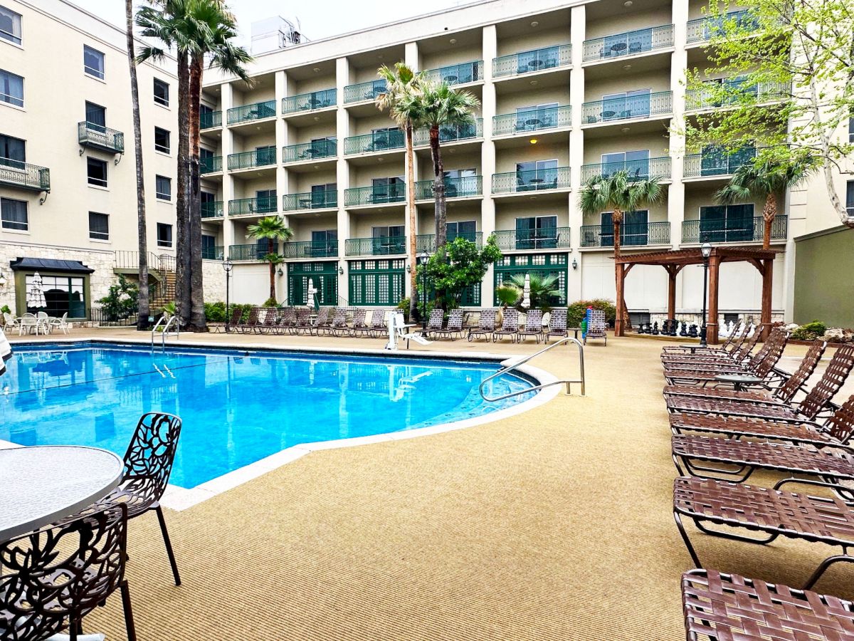 A hotel pool area with a large rectangular blue pool, surrounding lounge chairs, palms, and a multi-story beige building with balconies in the background.