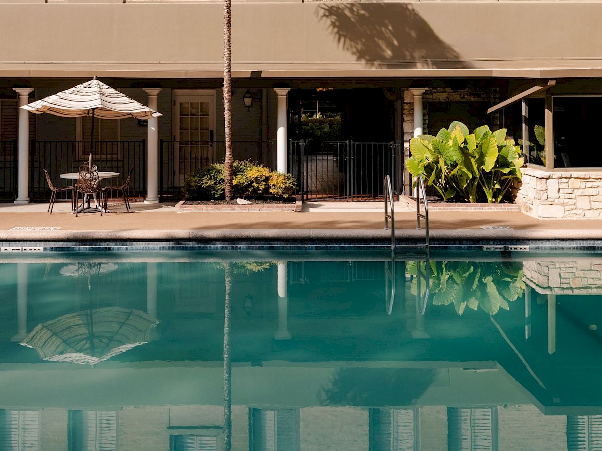 A quiet backyard pool with water reflections, white columns, patio seating, and leafy plants under a shaded umbrella.