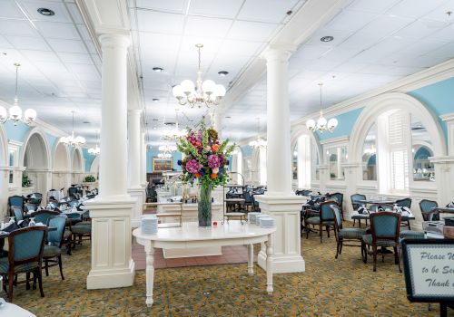 A bright, elegant dining hall with white columns, chandeliers, blue accents, and a central flower arrangement on a white table.
