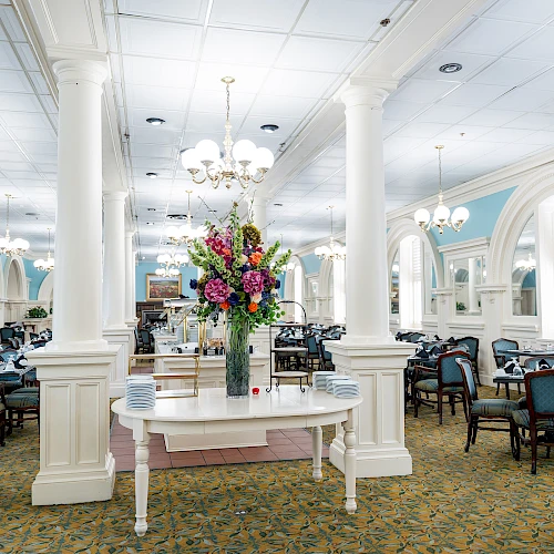 A bright, elegant dining hall with white columns, chandeliers, blue accents, and a central flower arrangement on a white table.