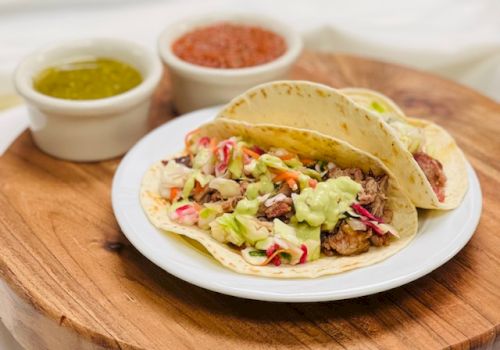 A plate of tacos with shredded meat, guacamole, and toppings on a wooden board, with two small bowls of salsa on the side.