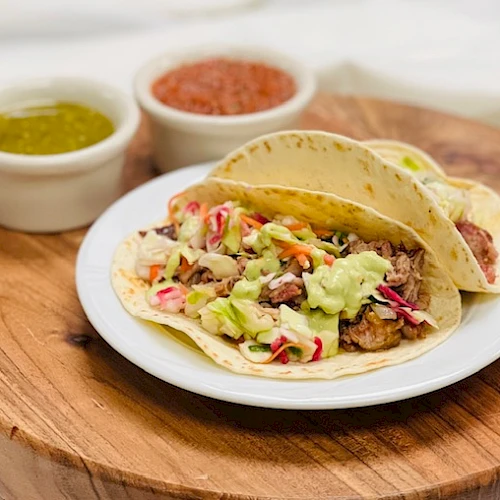A plate of tacos with shredded meat, guacamole, and toppings on a wooden board, with two small bowls of salsa on the side.