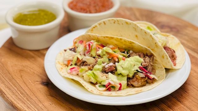 A plate of tacos with shredded meat, guacamole, and toppings on a wooden board, with two small bowls of salsa on the side.
