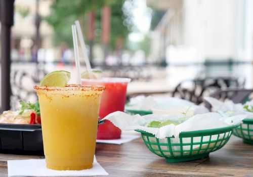 A sunny outdoor meal: a mango or orange smoothie with a spice rim, straw, lime wedge, plus bowls of food and chips on a communal table outdoors.