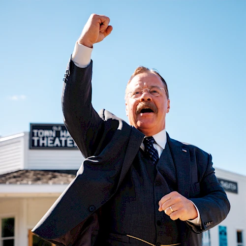 A jubilant man in a suit pumps his fist in the air outside a building labeled &ldquo;Theatre,&rdquo; celebrating a moment of triumph or success.