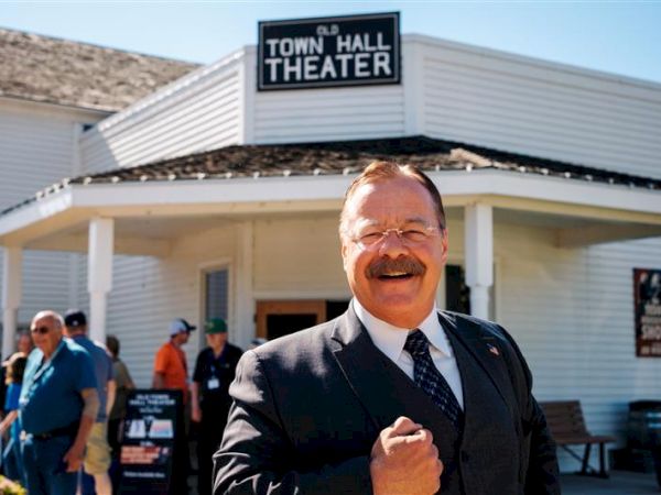 Feature: A smiling man in a suit foreground, outside a building labeled "Town Hall Theater," with people milling about in the background.