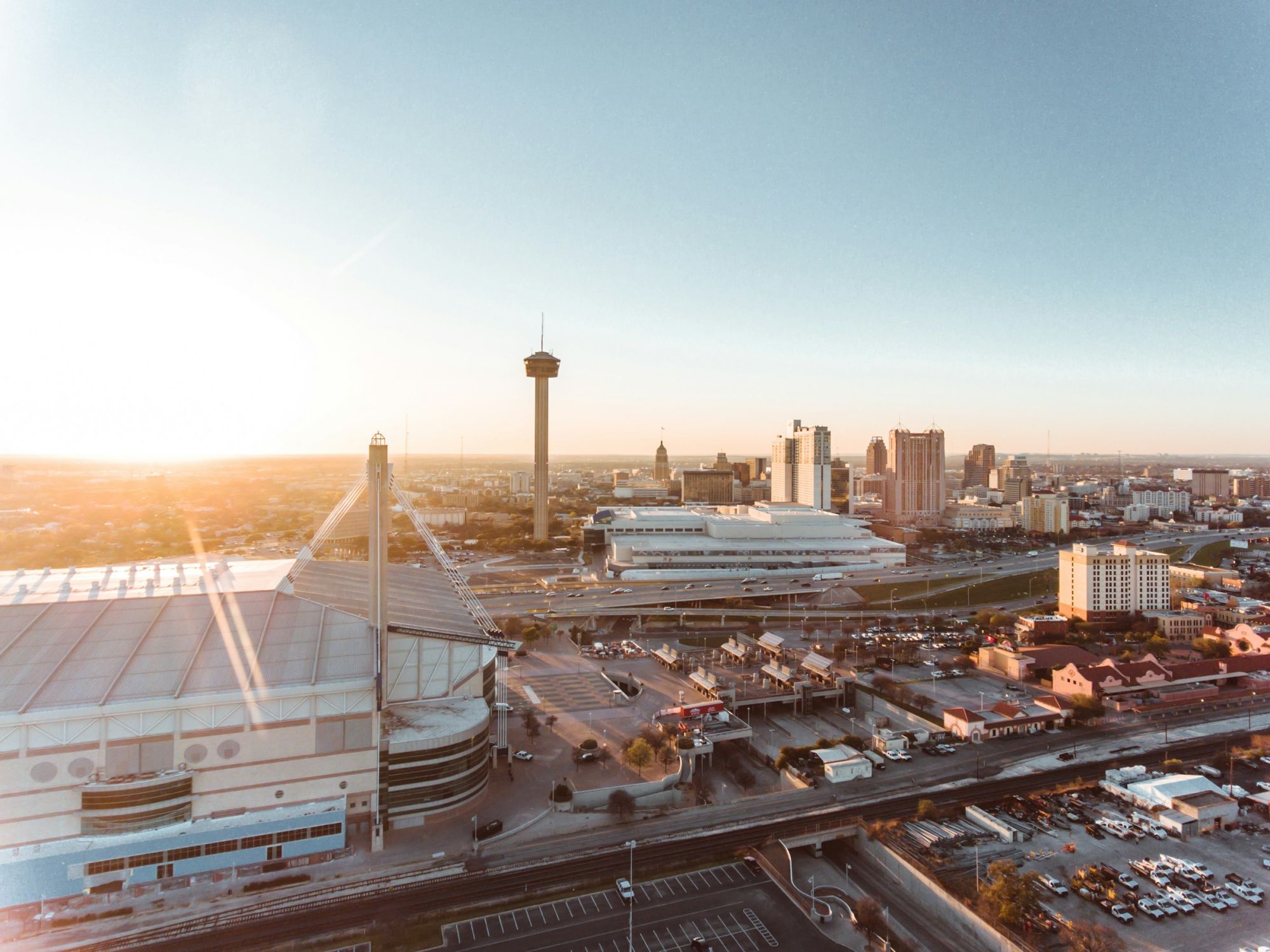 Aerial view of a city at sunrise with tall buildings, a network of roads, a stadium, and a traffic-filled riverfront area, bathed in warm light.
