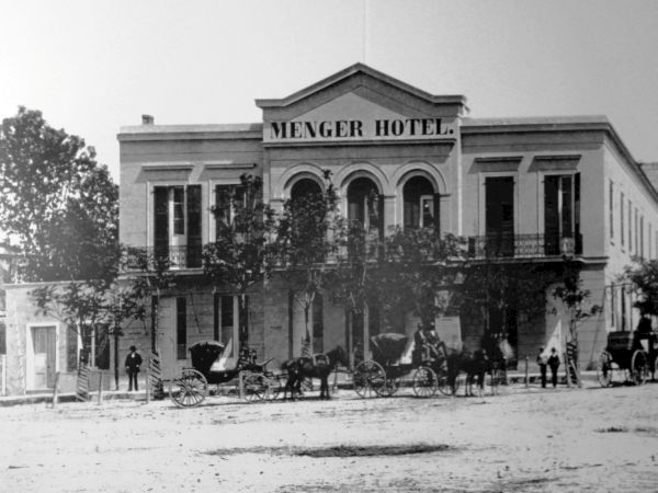 Old-fashioned street scene with a two-story hotel labeled &ldquo;Menger Hotel,&rdquo; horse-drawn carriages, and pedestrians in front.