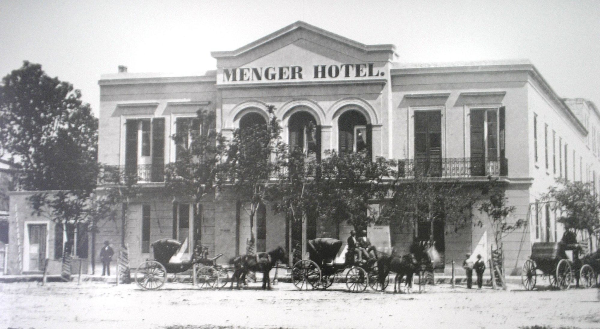 Old-fashioned hotel fa&ccedil;ade labeled &ldquo;Menger Hotel&rdquo;; horse-drawn carriages and people in vintage attire line the street in front.