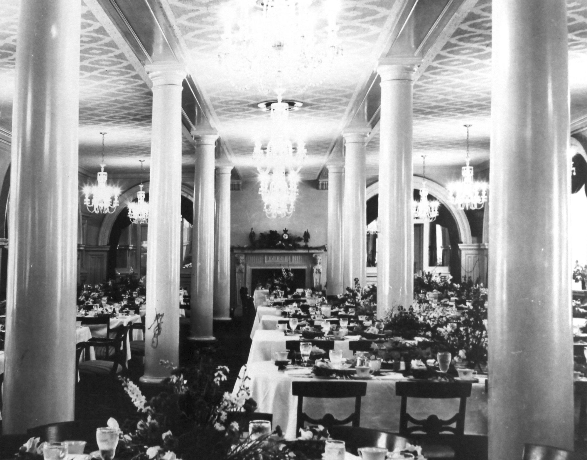 A formal dining hall with long tables, white tablecloths, chandeliers, and tall pillars, arranged for a banquet or event.