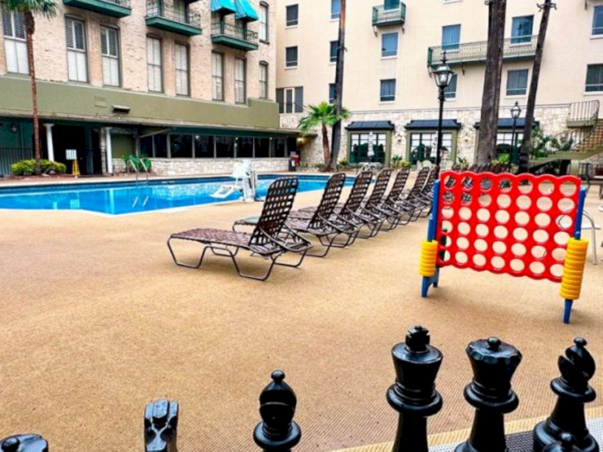 A resort-style pool area with lounge chairs, a bright red connect-four game, and a chessboard stopper in the foreground, sunny and inviting.