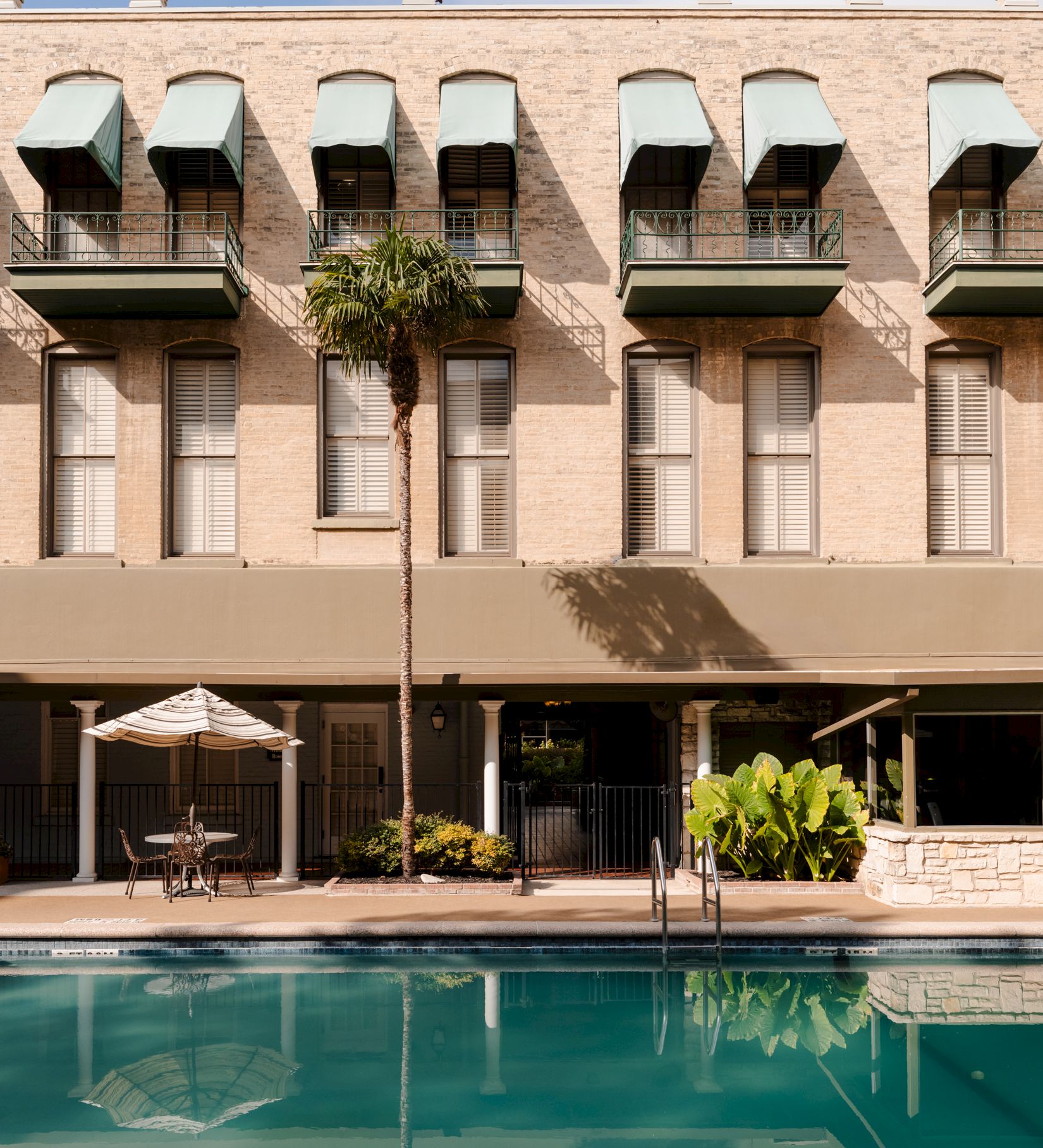 A stylish hotel facade with arched windows, balconies, and a ground-floor terrace; a calm outdoor pool reflects the building.