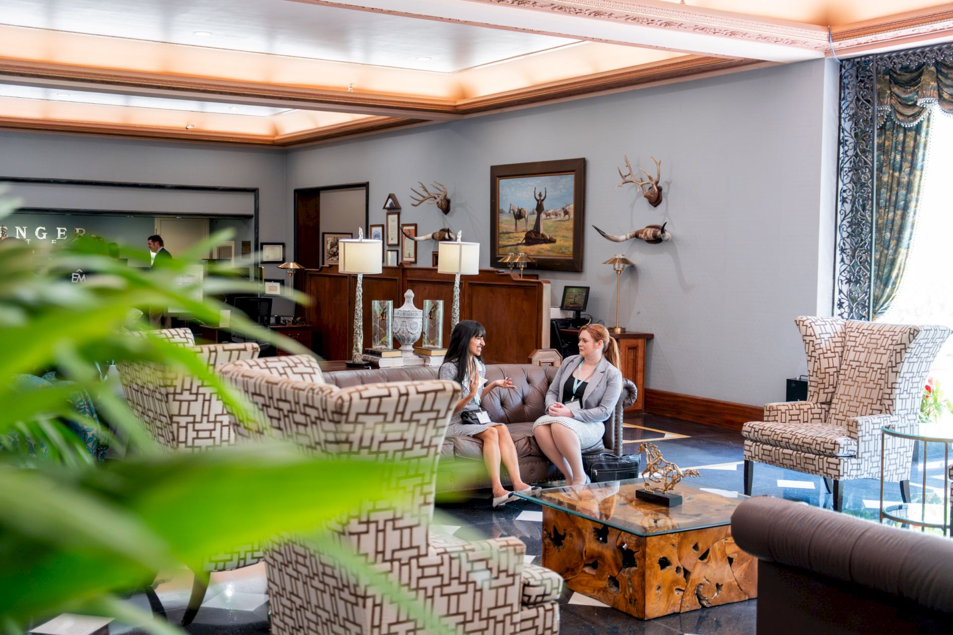 A hotel lobby with two women chatting near a reception desk, featuring wall art and mounted deer heads on the walls.
