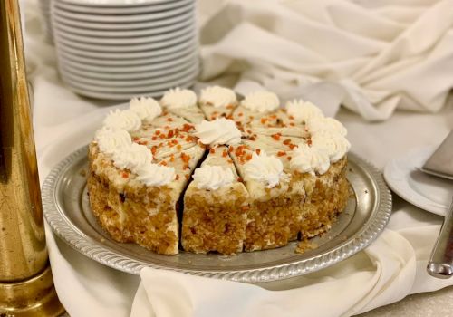 A frosted layered cake with white icing rosettes, sliced to reveal a nutty crumb, on a silver platter with a stack of plates in the background.