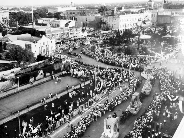 A vintage black-and-white city parade with a long marching procession down a broad street lined by spectators and early vehicles, crowds everywhere.