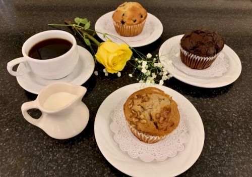 A coffee cup, a milk pitcher, a yellow rose with baby's breath, and four baked goods&mdash;three muffins on plates and one muffin on a doily, all on a dark countertop.