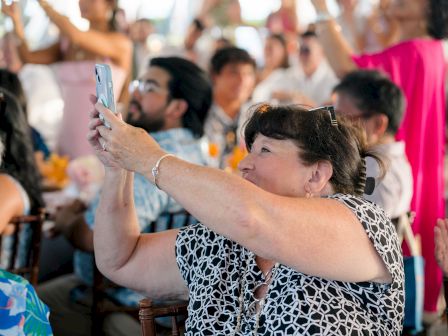 A woman takes a photo with her phone at a crowded event; people around her clap and cheer.