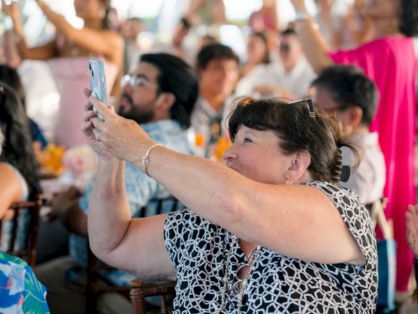 A woman takes a photo with her phone at a crowded event; people around her clap and cheer.