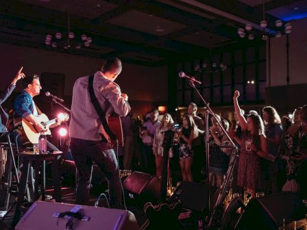 A live band performs on stage with guitars and a singer, crowd cheering with raised hands under stage lights.