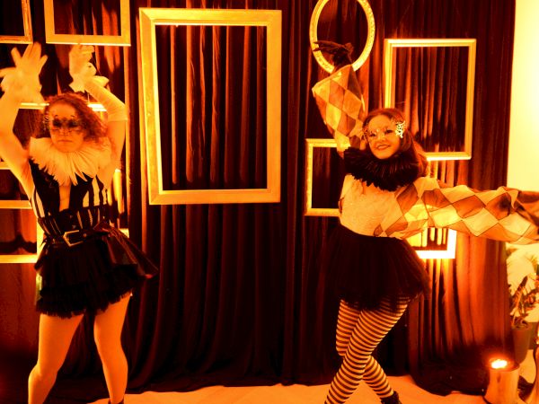 Two people dressed in festive, feathered outfits perform with props in a warm-lit stage setup featuring geometric frames and curtains.