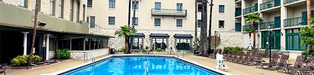 A hotel-style courtyard with a rectangular pool, lounge chairs, palm trees, and a beige, multi-story building surrounding the area.