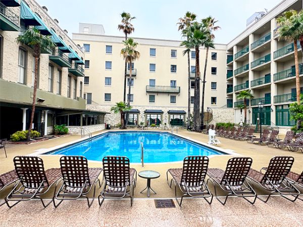 A motel-style courtyard pool scene with a bright blue rectangular pool, surrounded by lounge chairs and palm trees, flanked by beige buildings.