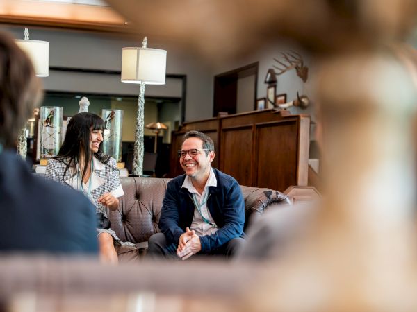 Three people chatting in a cozy lounge with warm lighting and wooden decor, smiling and seated on a sofa, casual vibes.