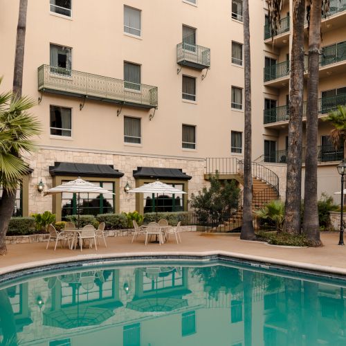 A rectangular pool in a courtyard of a beige apartment complex, with palm trees, patio tables, and shaded seating areas around it.