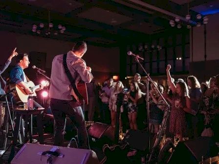 A live band performs on stage with guitars and a singer, while an enthusiastic crowd raises their hands and enjoys the concert.