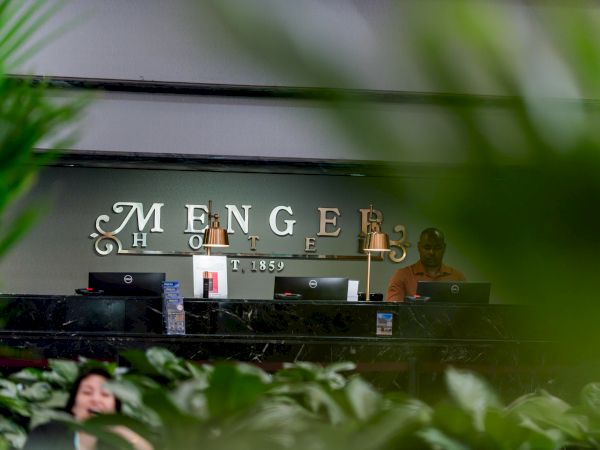 Hotel reception desk with computers, brass lamps, and a wall sign reading &ldquo;MENGE,&rdquo; seen through green leaves.