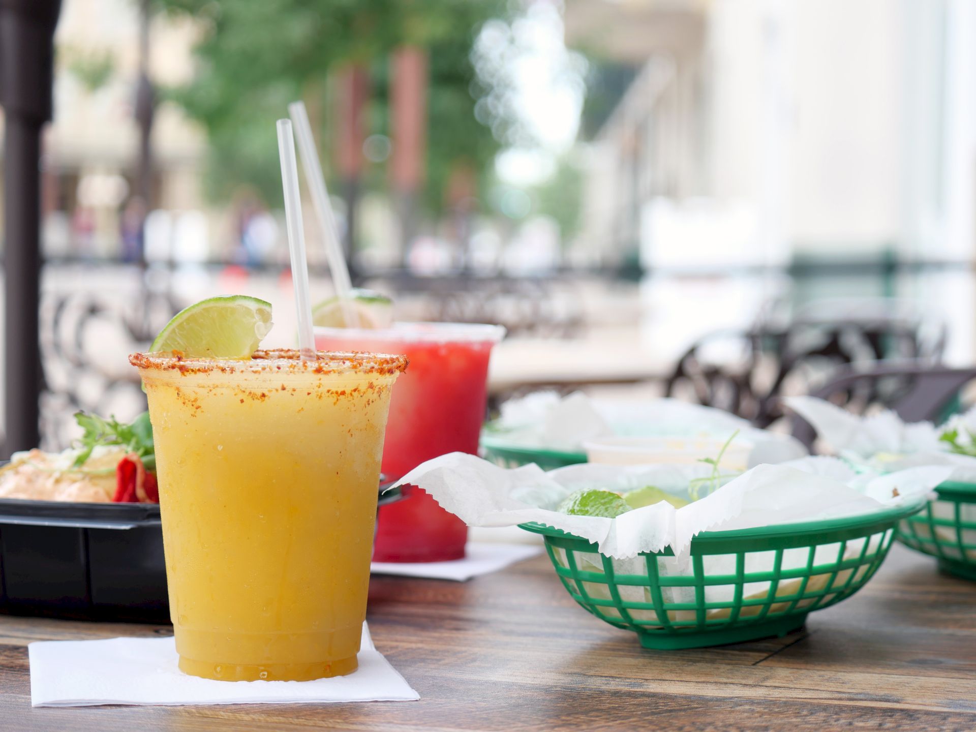A lime-rimmed drink with a lime slice and straw, with another red cocktail in the background on an outdoor patio.
