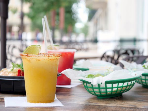 A lime-topped drink in a cup on an outdoor patio, with a second red cocktail behind and snacks nearby.