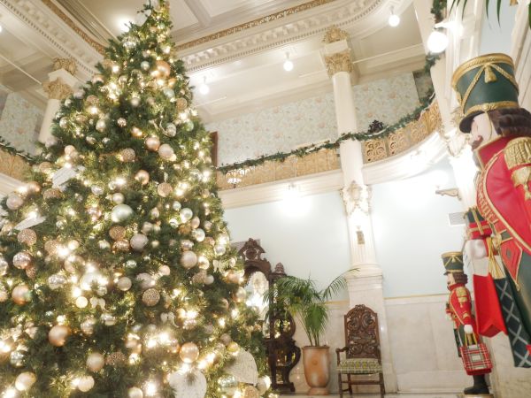 A huge decorated Christmas tree with warm lights fills an ornate hall, while a soldier nutcracker figure stands beside it.