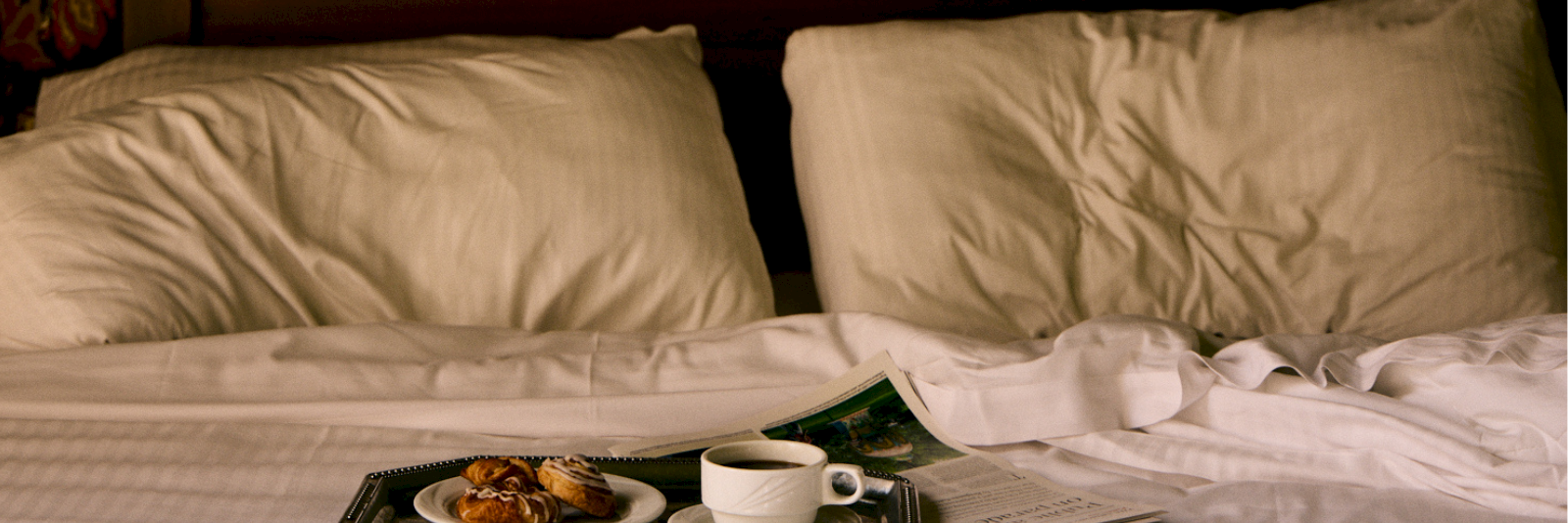 A made bed with pillows, a breakfast tray, a cup of coffee, and pastries spread out on top.