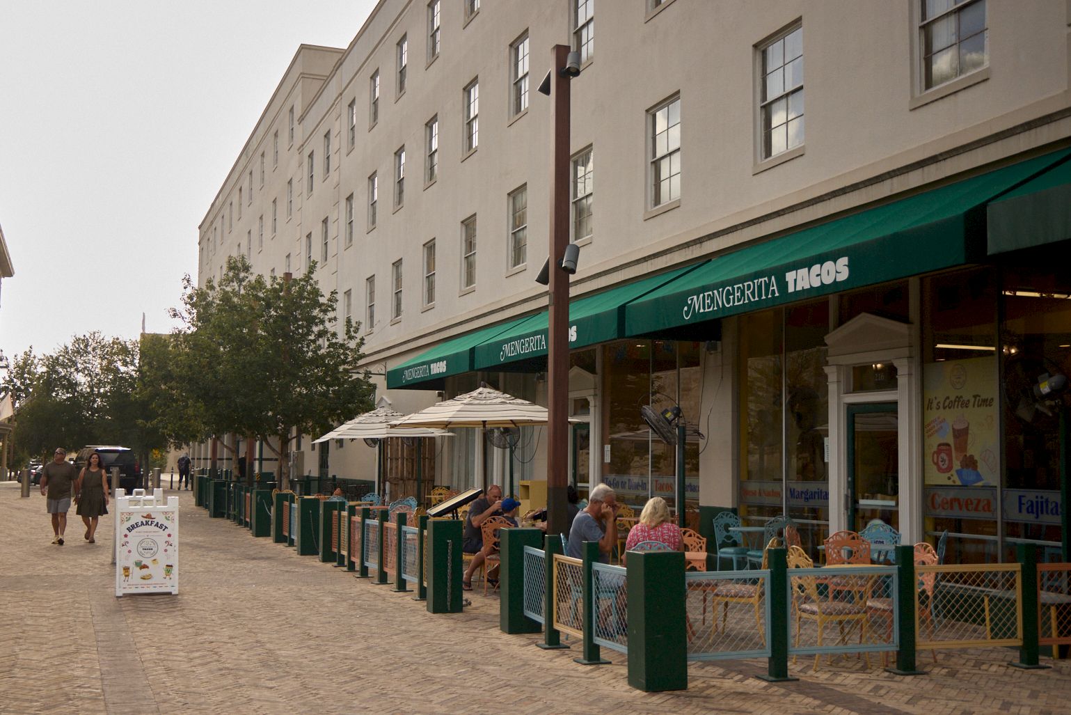 A sidewalk caf&eacute; strip with green awnings for Mengerita Tacos, people dining outside on a city street.