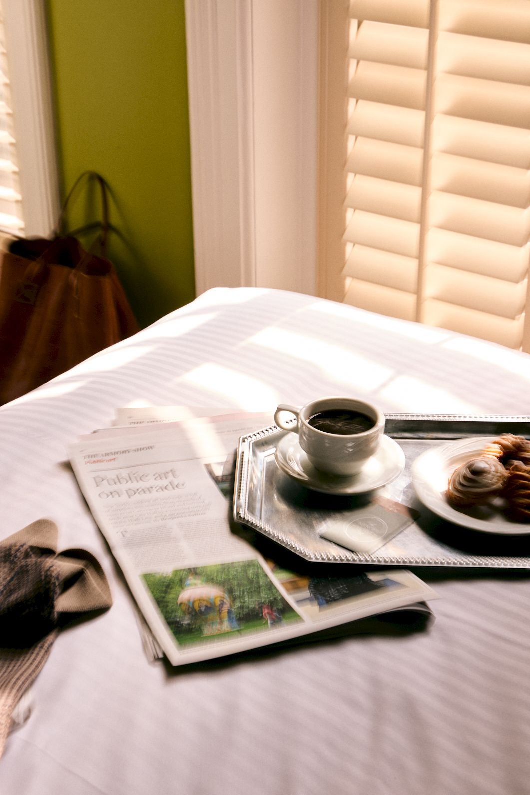 A sunny table with a cup of coffee, a tray, a croissant pastry, and a newspaper on a bed.